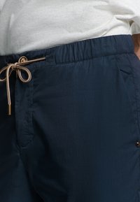 Marineblauwe zwembroek met een elastische tailleband, trekkoord en zijzakken. Gladde textuur met een subtiele glans. Accenten in goudtint.