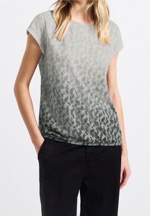 Kobieta w szarym topie w krótkim rękawie z nadrukiem w cętki, połączonym z czarnymi spodniami, stojąca z jedną ręką w kieszeni na białym tle.