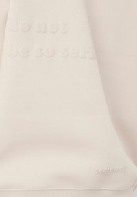 Sweat à la couleur beige clair avec un texte en relief : « ne sois pas si sérieux » et « sérieusement ». Texture lisse, col rond et ourlet côtelé.