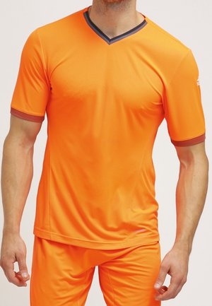 Mann trägt helloranges kurzärmeliges Sportshirt und passende Shorts, steht vor einem einfarbigen hellen Hintergrund.