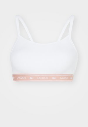 Bralette blanco con tirantes estrechos y una textura suave. Presenta una banda rosa clara con el logo "LACOSTE" y un emblema de cocodrilo.