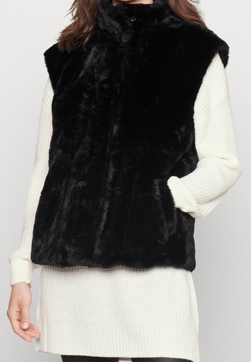 Veste noire sans manches en fausse fourrure portée par-dessus une robe-pull blanche en tricot à manches longues, les mains détendues sur les côtés.