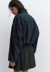 Ung kvinde med mørkt hår iført en løs mørk jakke og et plisseret skørt, set fra siden mod en ensartet lys baggrund.