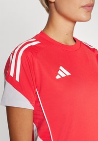 Rød idrettsskjorte med hvit tredelt mønster på skuldrene, hvite detaljer på ermene, og en hvit logo på forsiden. Laget av pustende stoff.