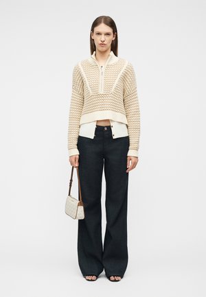 Γυναίκα που φοράει μπεζ πλεκτό κοντό πουλόβερ πάνω από λευκό πουκάμισο με κουμπιά, μαύρο παντελόνι με φαρδιά πόδια, κρατώντας μικρή λευκή τσάντα, στέκεται μπροστά σε λευκό φόντο.