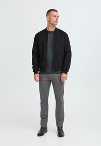 Musta samettinen bomber-takki tummanharmaan neuleen päällä, yhdistettynä slim-fit-harmaisiin housuihin ja mustiin nahkasaappaisiin. Yksinkertainen, minimalistinen muotoilu.
