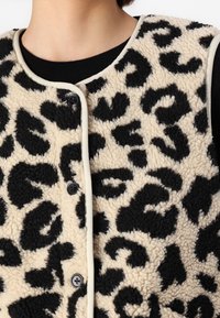 Närbild av en person som bär en beige och svart leopardmönstrad fluffig väst med svarta knappar över en svart skjorta.