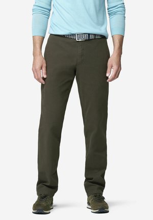 Mann trägt dunkel olivgrüne Hose, gemusterten Gürtel, hellblaues langärmeliges Hemd und grüne Sneakers und steht mit entspannten Armen.