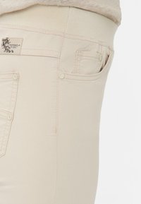 Beige Jeans mit einer glatten Textur, ausgestattet mit einem breiten Bund, einer kleinen Tasche und metallischen Akzenten. Etikett trägt die Aufschrift "Raphaela by Brax."