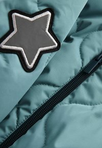 Un tissu en teal matelassé présente un patch étoile noir et gris ainsi qu'une fermeture éclair noire, mettant en avant une texture lisse et un design structuré.