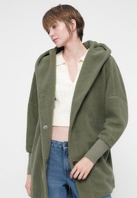 Obleka iz khaki zelene flis tkanine s kapuco, velikimi sprednjimi gumbi in rebrastimi manšetami. Oblikovana z udobnim krojem in mehkobo.