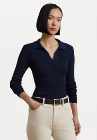 Pull en polo côtelé bleu marine avec un col en V, des manches longues et des poignets à revers ; associé à un pantalon clair et une ceinture noire avec une boucle dorée.