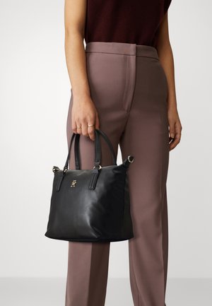 Femme tenant un sac à main en cuir noir avec logo doré, portant un pantalon taille haute mauve et un haut sans manches foncé, bagues visibles aux doigts.