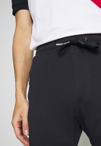 Pantaloni da jogging neri con vita regolabile tramite cordino, tasca laterale e etichetta Armani Exchange. Tessuto liscio, design aderente e cuciture pulite.