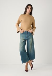 Pull en maille beige avec un col côtelé et des manches évasées, associé à un jean en denim à jambe large délavée. Modèles des escarpins bordeaux pointus.