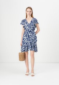 Žena v modro-bielych kvetinových šatách na zábal, držiaca tkanú kabelku, stojaca pred jednoduchým bielym pozadím a v bielych sandáloch.