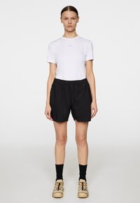 Vit kortärmad skjorta med en liten logotyp, kombinerad med svarta shorts med elastisk midja. Bärs med svarta strumpor och beiga mönstrade skor.