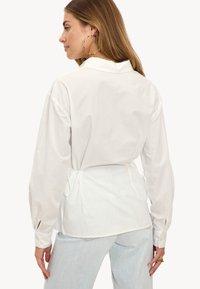 Blusa de algodón blanca con mangas largas, cuello, lazos en la espalda y aberturas laterales. Combinada con jeans de color azul claro.