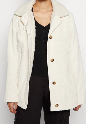Person iført en off-white kordfløjl jakke med for i shearling og brune knapper over en sort ribbet knappet top og sorte bukser.