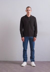 Moški stoji oblečen v črno dolgimi rokavi s kragnico, modre kavbojske hlače in bele superge pred enobarvno svetlo sivo steno.