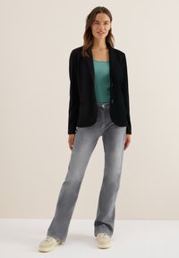 Femme debout portant un blazer noir, un haut canard, un jean gris et des baskets beiges, devant un fond beige clair.