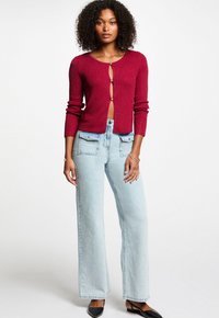 Cardigan corto a coste bordeaux con chiusura frontale a bottoni, abbinato a jeans a gamba larga azzurri con tasche frontali e finitura scolorita.