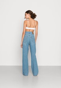 Jean de cintura alta azul claro com perna reta, apresentando um patch de couro castanho e dois bolsos traseiros. Modelo a usar um bralette branco.