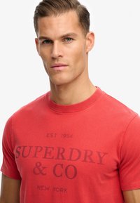 Czerwona bawełniana koszulka z okrągłym dekoltem, krótkimi rękawami oraz nadrukiem z przodu z napisem "SUPERDRY & CO NEW YORK" w ciemnym kolorze.