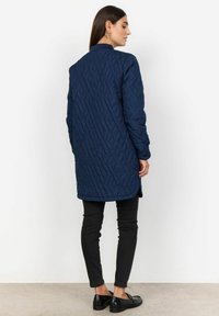 Marinblå vadderad lång jacka med rak skärning, strukturerad design, rundad nederkant och sidofickor, kombinerad med svarta skinny jeans och loafers.