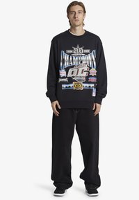 Black sweatshirt with graphic prints, including "Champions" and various logos. Paired with loose black pants and black sneakers.