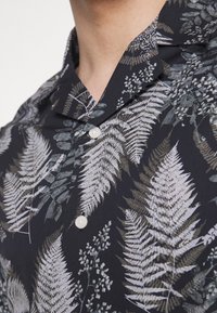 Donker shirt met een varen- en bladdessin in grijs- en groentinten, met een scherpe kraag en twee witte knopen bij de hals.