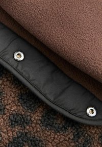 Smeđa flis tkanina složena preko crnog materijala, s detaljem srebrnog dugmeta na kopčanje i teksturiranom smeškom i crnom uzorkom ispod.