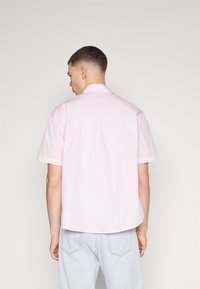 Camisa de manga corta en algodón rosa claro. Cuenta con un cuello clásico, detalle de yugo en la espalda y dobladillo ligeramente redondeado. Combinada con pantalones de mezclilla claros.