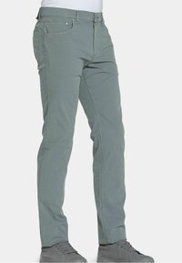 Muž v svetle šedých volných kalhotech a šedých teniskách, stojící na bílém pozadí.