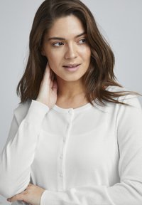 Cardigan bianco lavorato a maglia con maniche lunghe e colletto rotondo, caratterizzato da una fila di bottoni sul davanti. Texture morbida e design semplice.