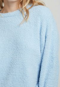 Pull en maille texturée bleu clair en tissu doux et moelleux. Il présente un col rond et des manches longues, offrant une coupe décontractée.