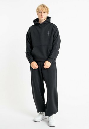 Czarna bluza z kapturem i przednią kieszenią, zestawiona z luźnymi, czarnymi spodniami. Materiał wydaje się miękki, z luźnym krojem i dzianinowymi ściągaczami.