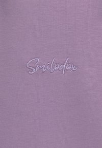 Lila Baumwollstoff mit glatter Textur, auf dem das gestickte Wort "Smiledox" in weißen Fäden mittig auf dem Material platziert ist.