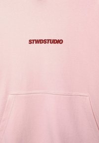 Pink sweatshirt with front pocket, featuring a maroon "STWDSTUDIO" logo. Fabric appears soft with a smooth texture and a relaxed fit.