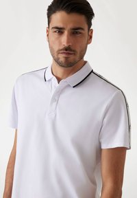 Polo blanco con cuello estándar, con ribetes negros en el cuello y las mangas. Tejido suave y transpirable; con tapeta de dos botones.