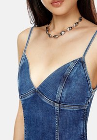 Kvinna i en blå denimtop med spaghettiband och ett chunky silverpärlhalsband, sedd från axlarna till mitten av bröstet.