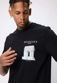 Homme avec des dreadlocks portant un t-shirt noir illustré d'un texte blanc de la Renaissance et d'une illustration détaillée d'un monument en arc classique.