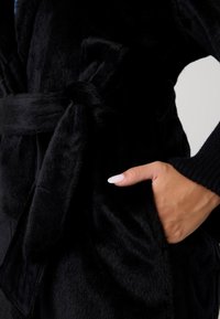 Schwarzer Morgenmantel aus Kunstpelz mit Bindegürtel, der eine weiche Textur und eine Seitentasche besitzt. Der Ärmel ist mit einem Rippstrick-Akzent versehen.