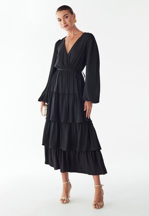 Schwarzes, fließendes Maxikleid mit langen Ärmeln, tiefem V-Ausschnitt und abgestuften Rüschen. Weicher Stoff mit elastischem Taillenband und lockerer Passform. Goldene High Heels als Accessoire.