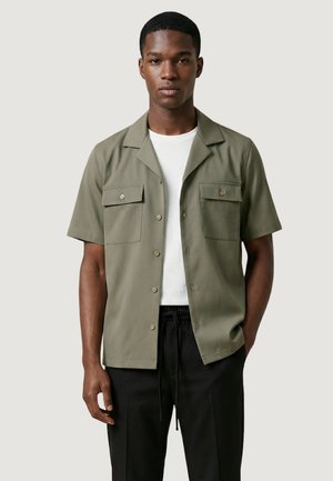 Jeune homme portant une chemise boutonnée à manches courtes de couleur vert olive avec deux poches poitrine, par-dessus un t-shirt blanc et un pantalon noir.