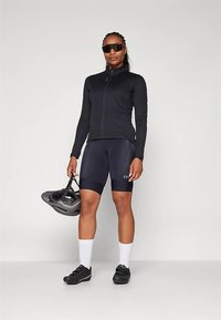 Black cycling jacket and navy shorts, holding a helmet. Sunglasses, white socks, and black shoes complete the activewear look.