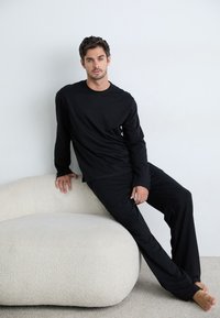 Ensemble de chemise à manches longues noires et pantalon, fabriqué en tissu doux, avec une coupe décontractée. Le modèle est assis sur un canapé texturé crème.