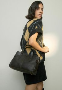 Woman wearing a black and tan patterned sleeveless sweater and black skirt, carrying a large black leather handbag over her shoulder.