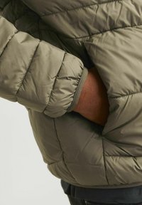Olijfgroen pufferjack met een gequilt patroon en elastische manchetten. De manchetten hebben een contrasterende bies. Een zak is zichtbaar aan de zijkant.