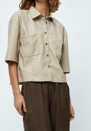 Kobieta w beżowej koszuli z krótkim rękawem z guzikami i kieszeniami z przodu oraz luźnych ciemnobrązowych spodniach, stojąca na tle jednolitego tła.